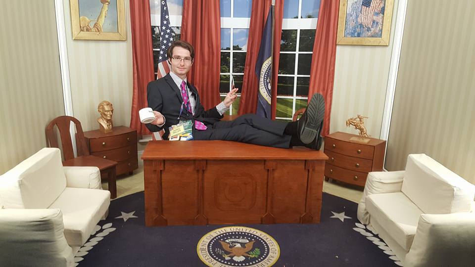 Society President Miller sitting in a mini Oval Office holding a coffee cup and American flag